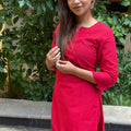 Three-quarter front view of a woman wearing a red handloom cotton kurta set near green foliage.