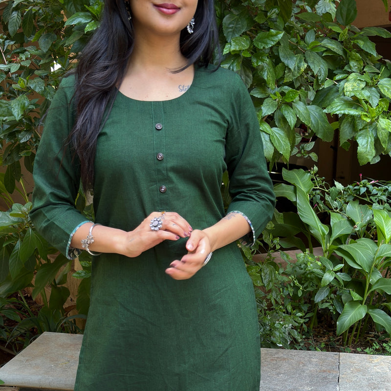 Full height portrait of the woman in the dark green cotton kurta set standing on a stone pathway outside.