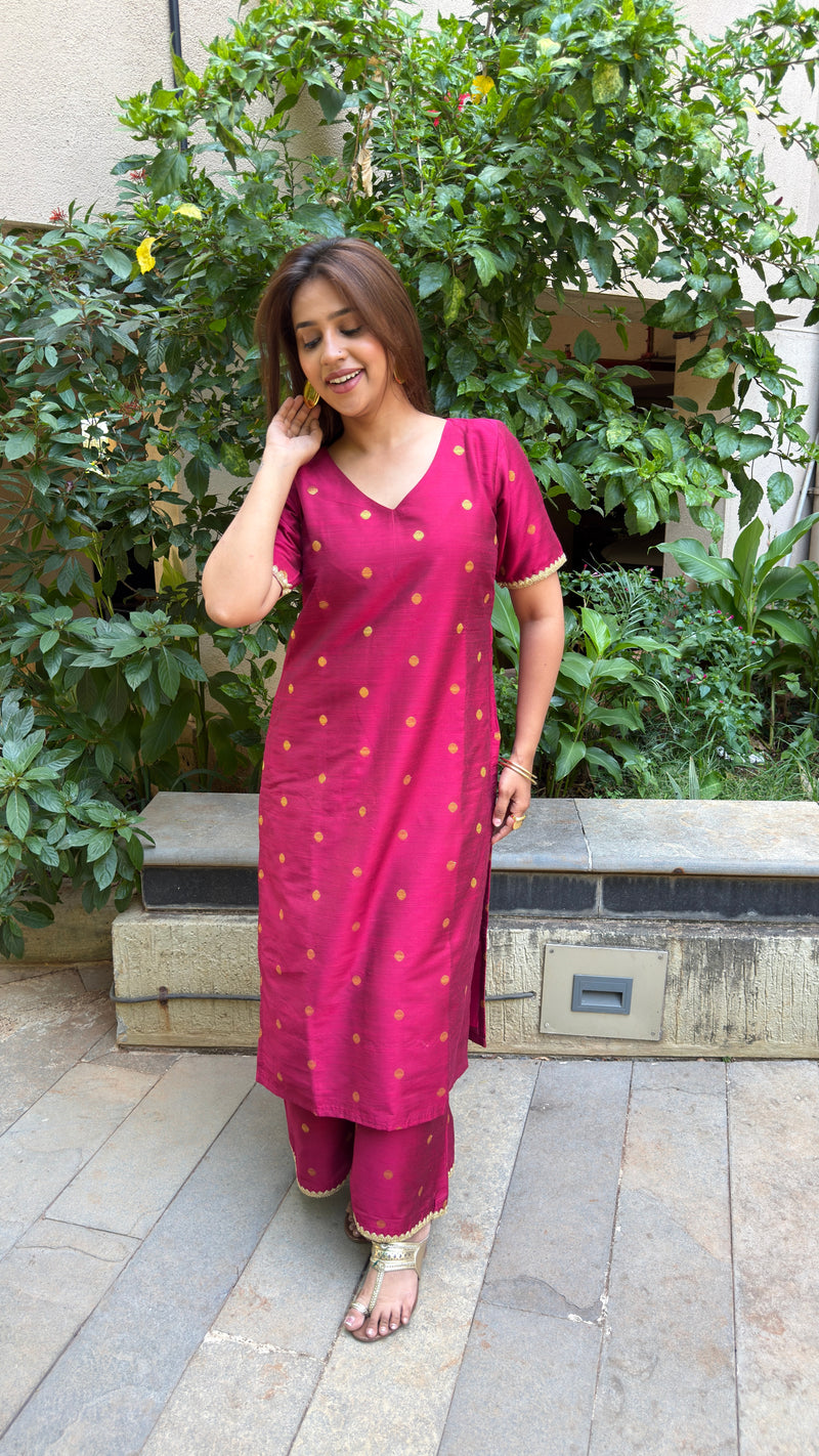 A woman standing in a garden wearing a magenta polka dot slub silk kurta set paired with wide trousers.